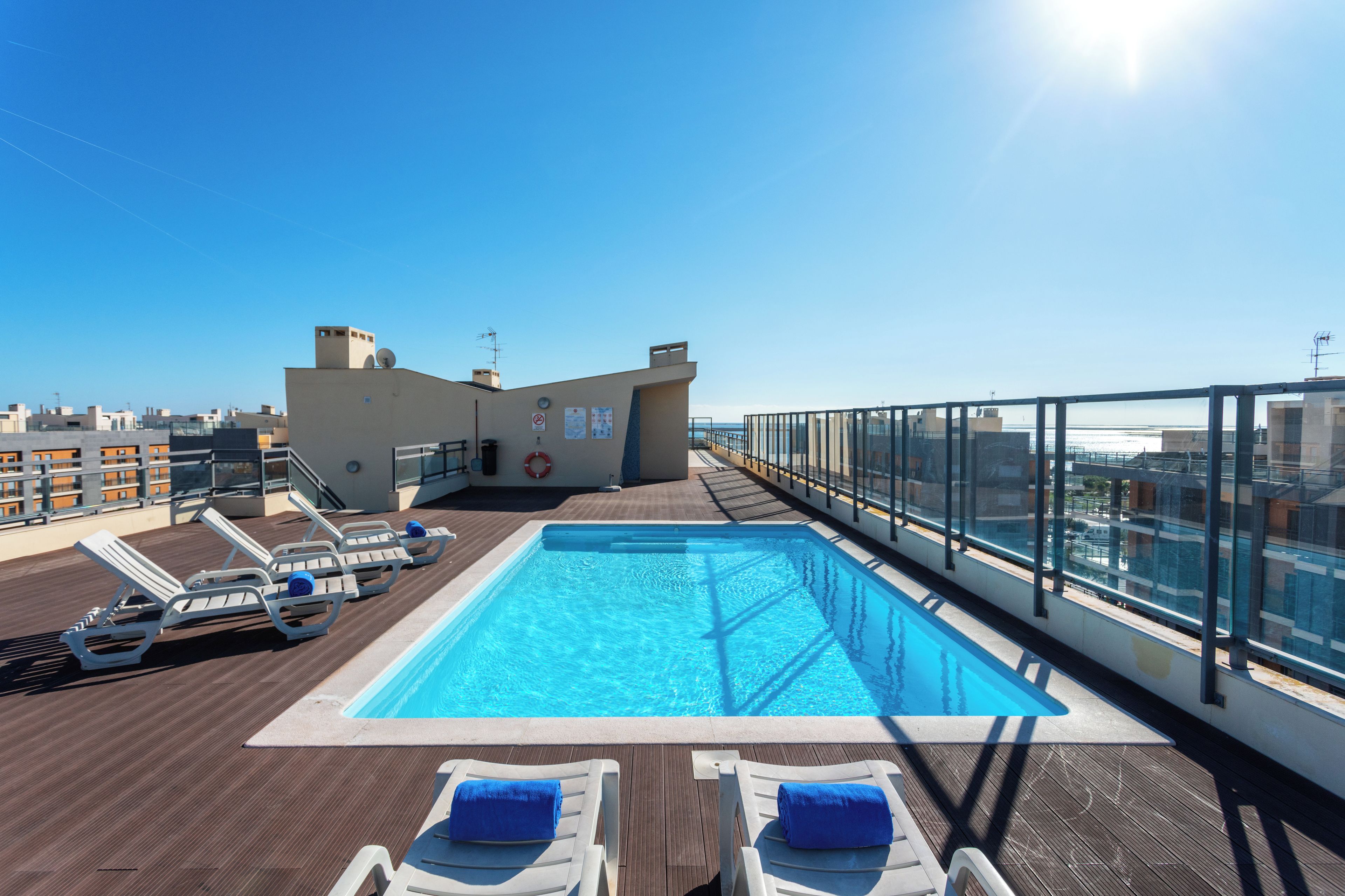 A rooftop pool with clear blue water, surrounded by lounge chairs and a glass railing overlooking a cityscape.
