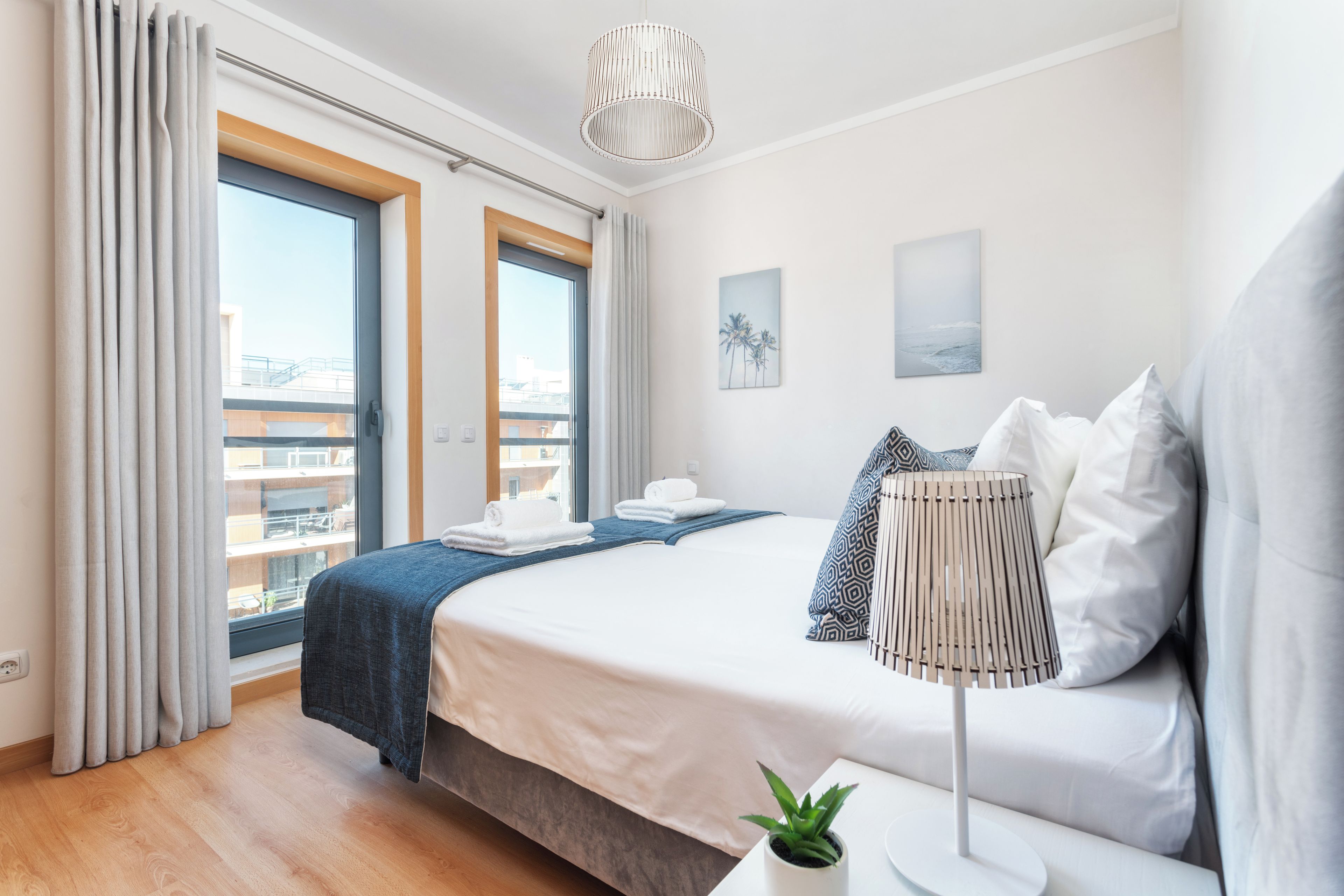 A modern hotel room with a large bed, a bedside table with a lamp, a view of the city through the window, and a balcony with a railing.
