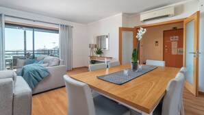 Una moderna camera d'albergo con un tavolo da pranzo in legno, sedie bianche, un divano e un'ampia finestra con vista sulla città.