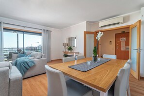 Een moderne hotelkamer met een houten eettafel, witte stoelen, een bank en een groot raam met uitzicht over de stad.