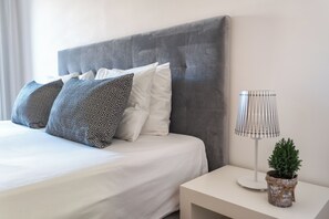 A neatly made bed with patterned pillows, a grey upholstered headboard, a bedside lamp, and a small potted plant on a bedside table. - Privilege 2-Bedroom Apartment with Rooftop Pool Terrace, Village Marina - Olhão (Olhão)
