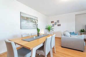 A modern living room with a wooden dining table, white chairs, a grey sofa, and a painting on the wall. - Privilege 2-Bedroom Apartment with Rooftop Pool Terrace, Village Marina - Olhão (Olhão)
