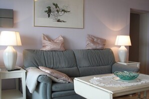 A living room with a grey sofa, two white side tables, a white coffee table with a decorative bowl, and a framed floral artwork on the wall. - Apartment CuxNautic414 (Cuxhaven)