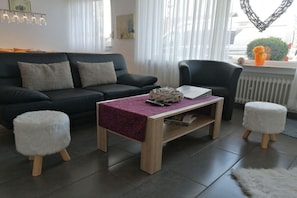 A living room with a black sofa, a wooden coffee table, two white poufs, and a chair. - "To the Moselle View" (Reil)