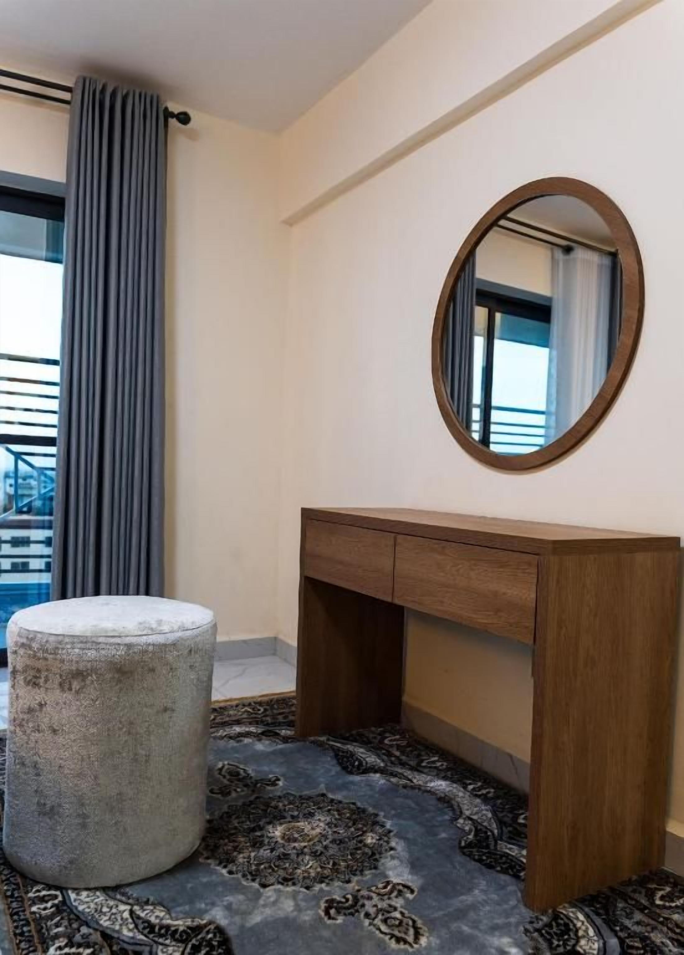 Una habitación de hotel con un escritorio de madera, un espejo redondo, un otoman gris y una alfombra estampada.