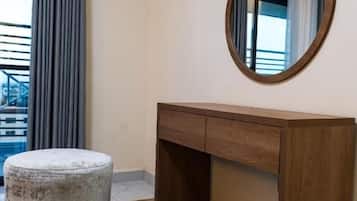 Una habitación de hotel con un escritorio de madera, un espejo redondo, un otoman gris y una alfombra estampada.
