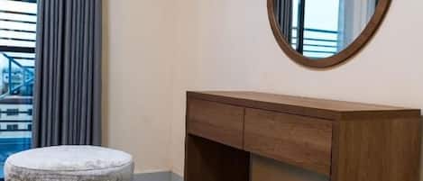 Una habitación de hotel con un escritorio de madera, un espejo redondo, un otoman gris y una alfombra estampada.