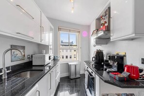 Eine moderne Küche mit weißen Schränken, einer schwarzen Arbeitsplatte, einer Kaffeemaschine und einem Fenster mit Blick auf Gebäude.