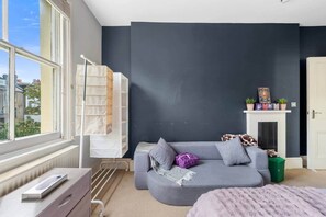 Ein modernes Schlafzimmer mit grauem Sofa, einem kleinen Tisch, einer Heizung, einem Fenster mit Blick auf Gebäude und einem Kamin mit Dekoration.