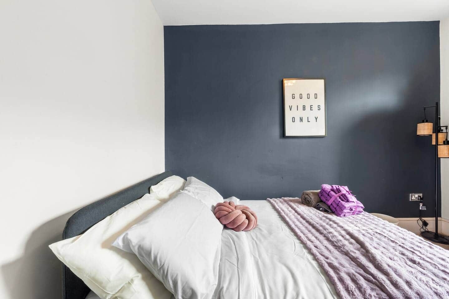 A bedroom with a bed, pillows, a blanket, a wall-mounted light fixture, and a framed motivational quote.