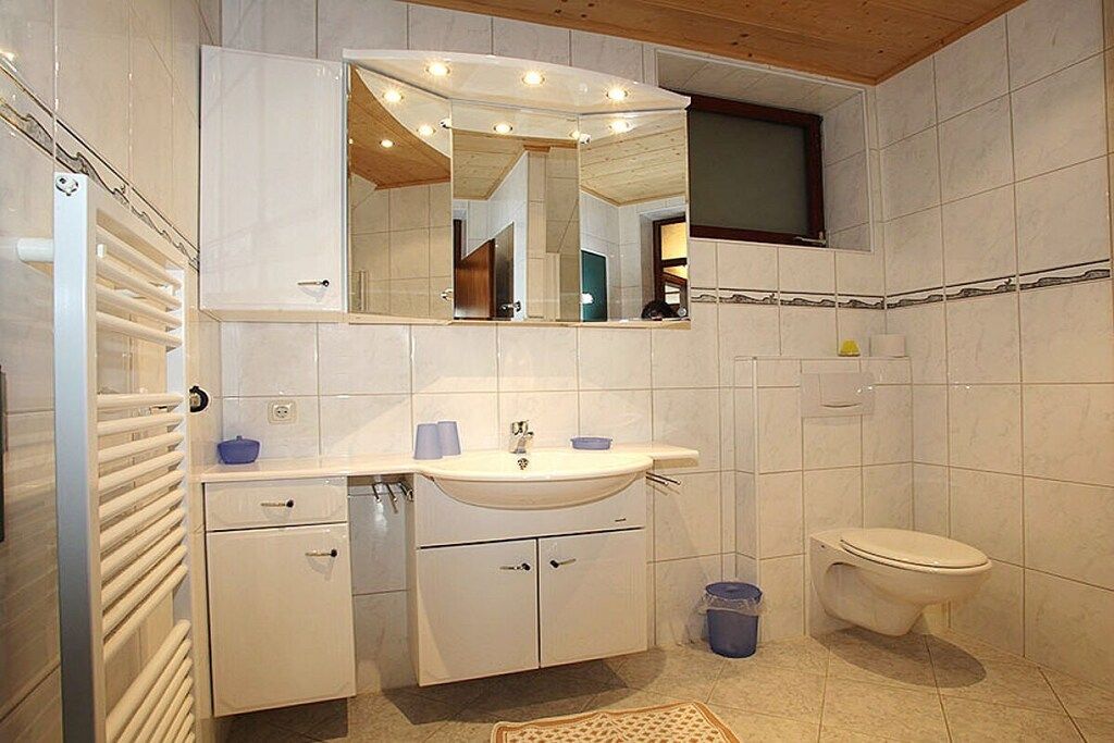 A bathroom with a white sink, mirror, and toilet. There is a towel rack and a small trash bin.