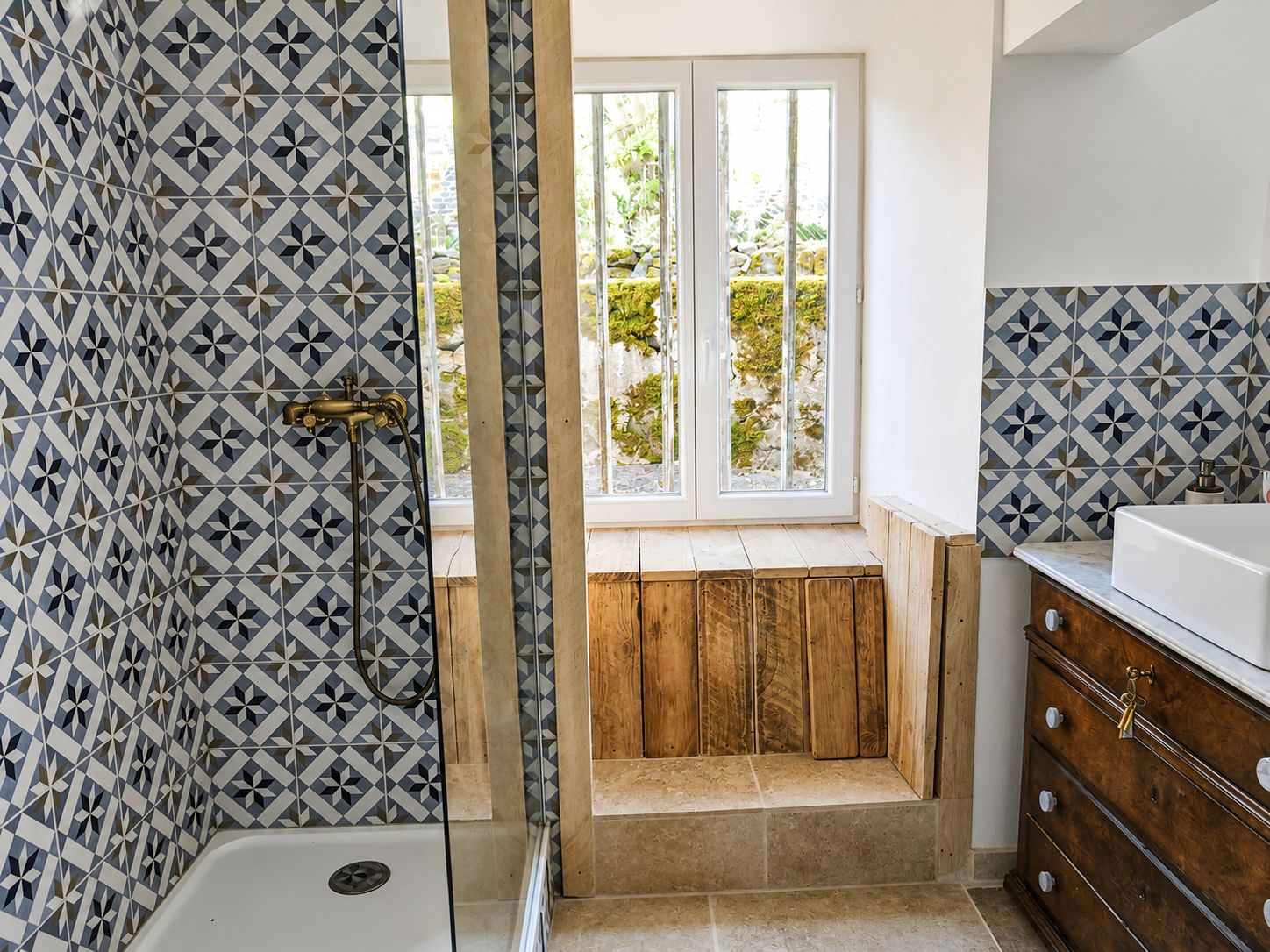 A bathroom with a geometric tile pattern, a shower, a wooden bench, and a view of greenery outside.