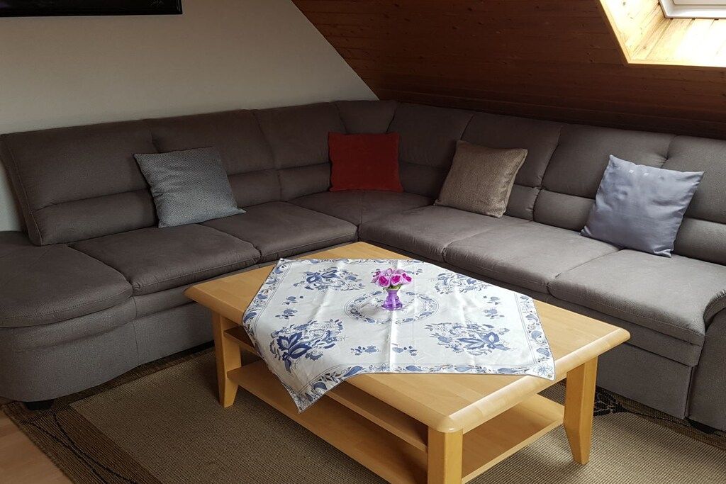 A corner living room with a grey sectional sofa, a wooden coffee table, and a patterned tablecloth.