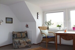 A room with a sloped ceiling, a patterned sofa, a wicker chair, a wooden table, and a large window with plants. - Holiday apartment with 1 bedroom (Osteel)