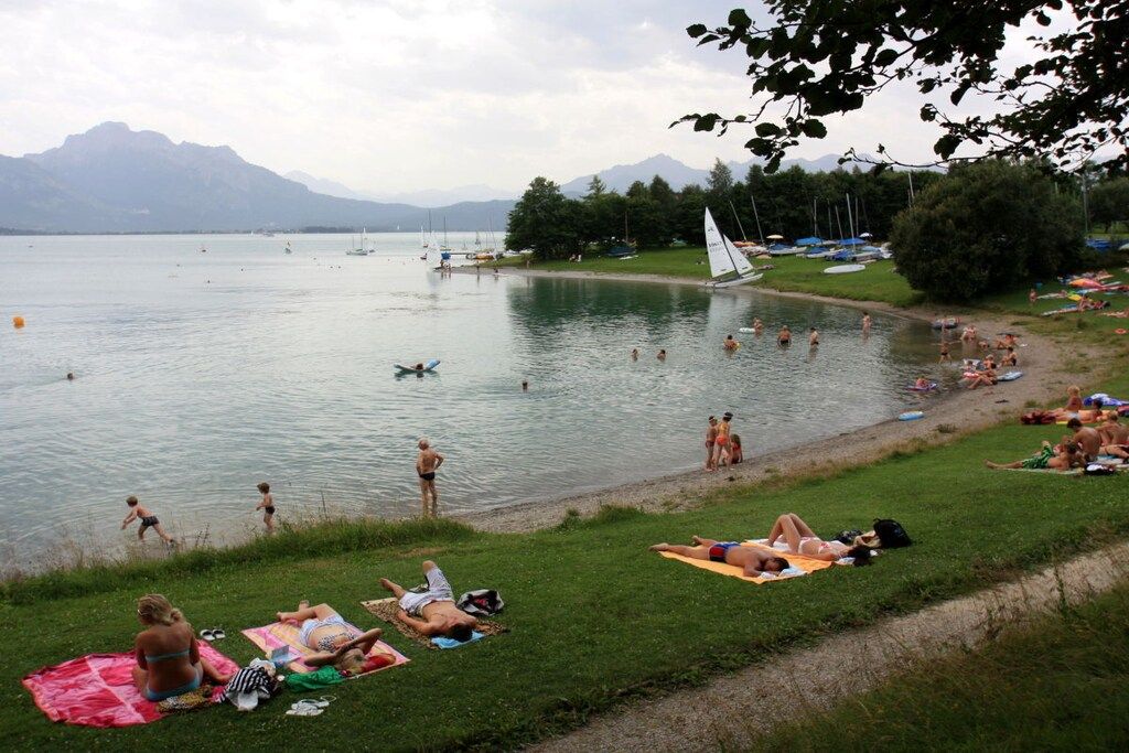 Une scène au bord d’un lac, où l’on voit des gens se prélasser au soleil et nager, des bateaux sur l’eau et un voilier amarré près du rivage.