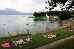 A lakeside scene with people sunbathing and swimming, boats on the water, and a sailboat docked near the shore. - Holiday apartment with 1 bedroom (Roßhaupten)