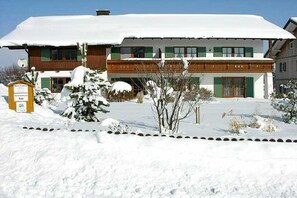 A two-story building with a snow-covered roof, a wooden facade, and a signboard in the foreground. - Holiday apartment with 1 bedroom (Roßhaupten)