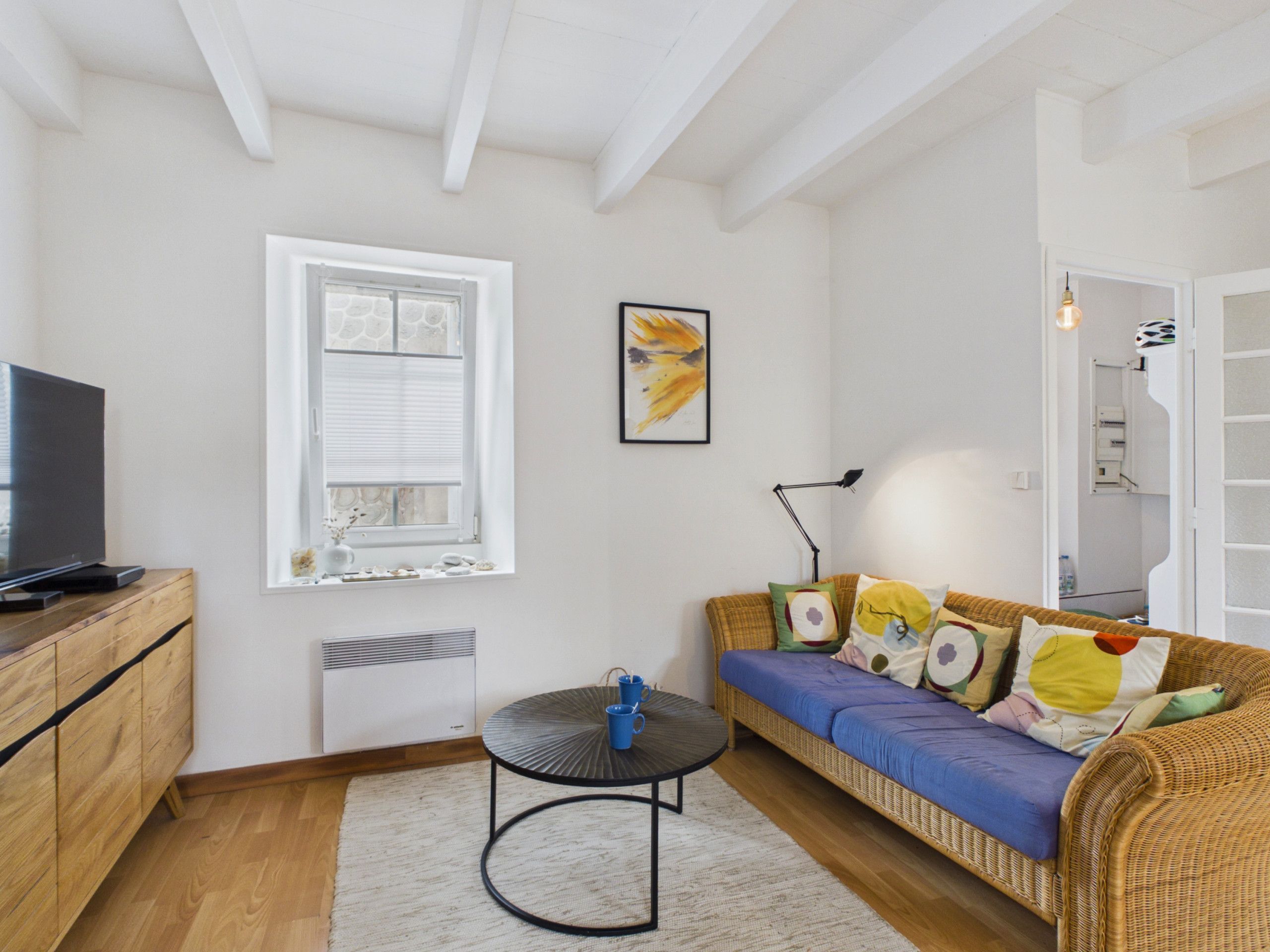 Una sala de estar con sofá, televisión sobre un mueble de madera, ventana con persianas, un cuadro en la pared y una lámpara.
