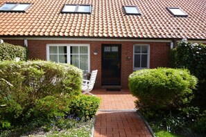 A brick house with a tiled roof, a front door, and a small garden with a pathway leading to a white chair. - Holiday apartment with 2 bedrooms (Greetsiel)