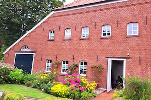 A red brick building with a large garage door, multiple windows, and a garden with various flowers. - Holiday apartment with 2 bedrooms (Greetsiel)