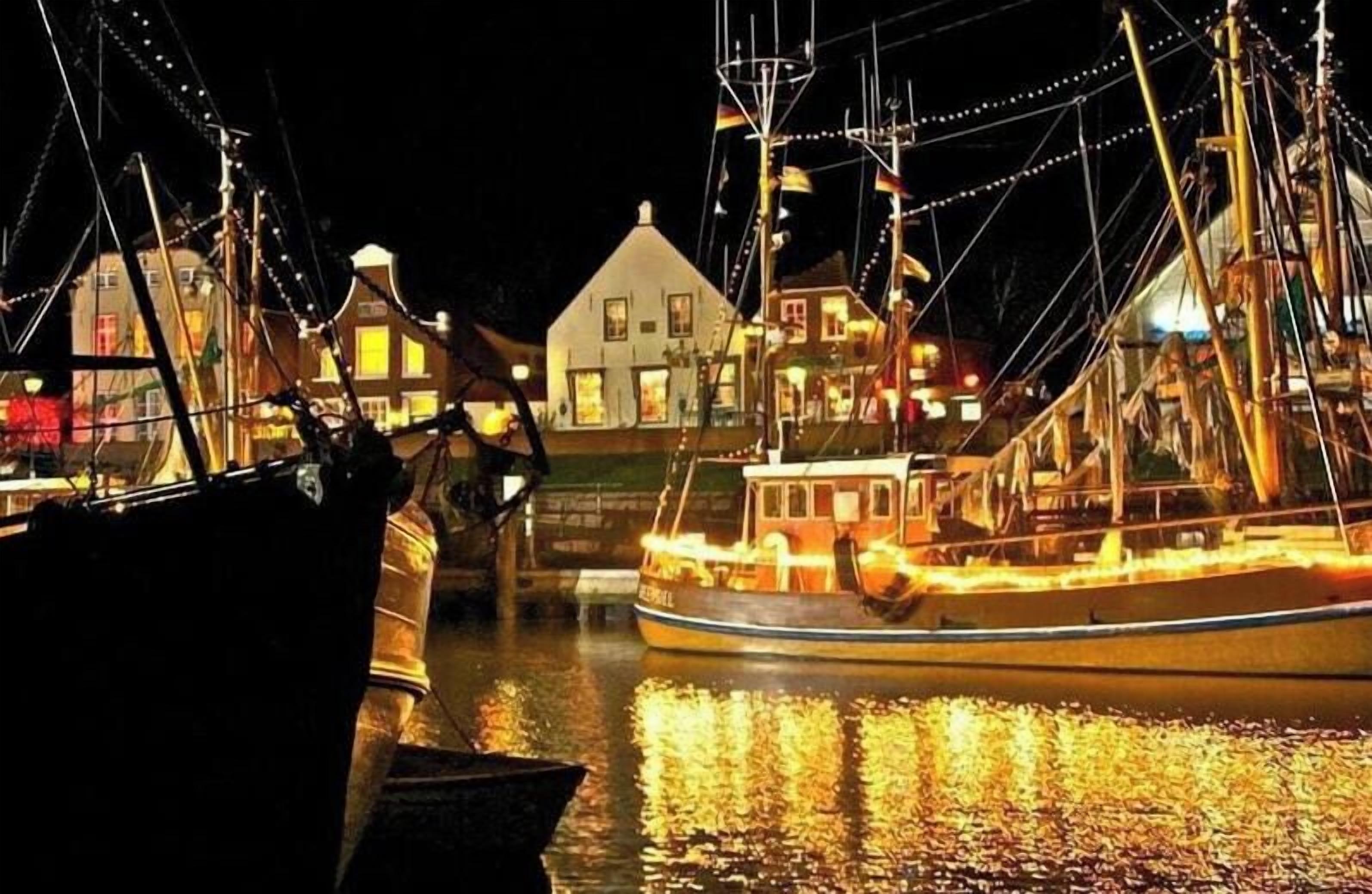 Cena noturna de um porto com barcos atracados, um edifício iluminado e uma superfície de água calma refletindo as luzes.