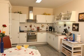 Een kleine keuken met witte kastjes, een fornuis en een koffiezetapparaat. Er staat een eettafeltje met een witte tafelkleed en twee glazen.