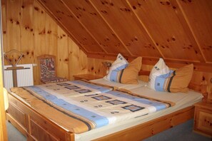 Ein Zimmer mit Holzboden und schrägem Dach, ein Bett mit gemusterten Textilien, ein Sessel und ein kleiner Tisch.