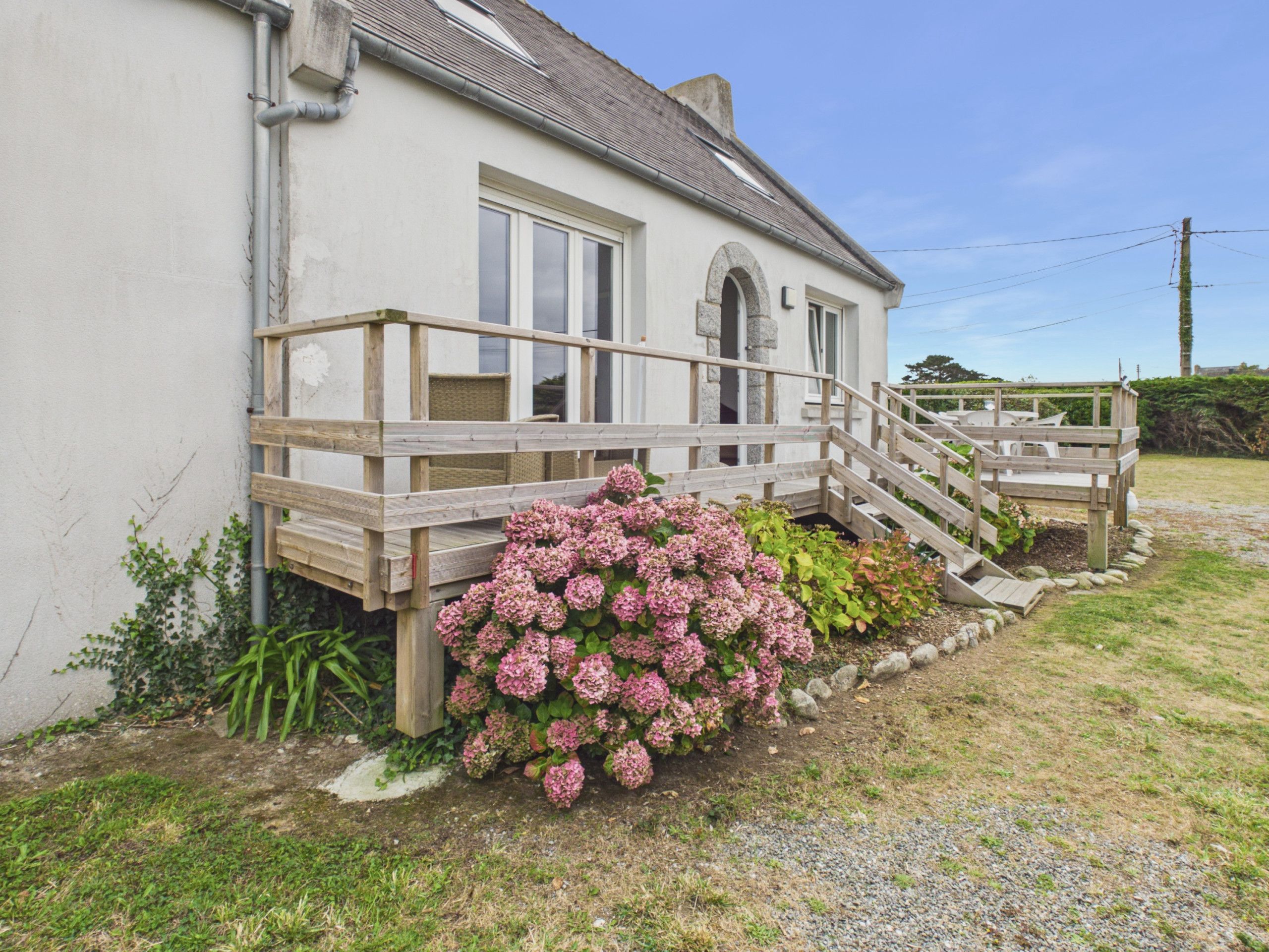 Une maison dotée d’une terrasse en bois, d’une arche en pierre et d’un jardin où poussent des fleurs roses.