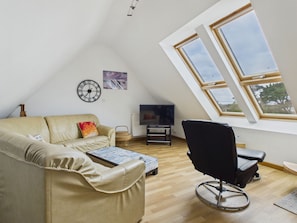 A room with a sloped ceiling, a sofa, a black chair, a television, and a window.
