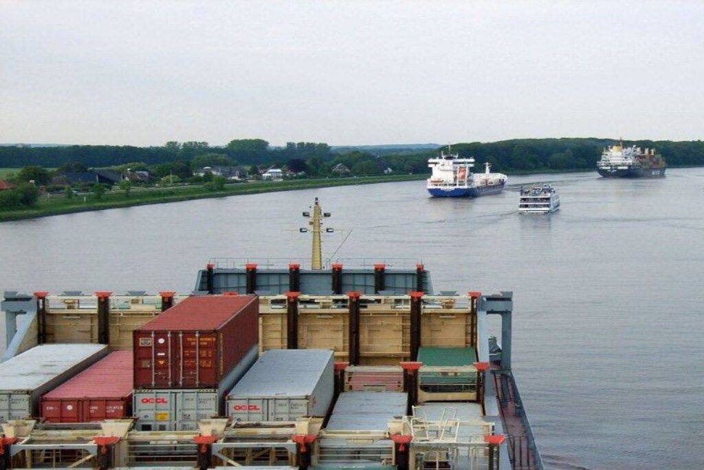 Ein Frachtschiff mit Containern und ein Schlepper im Vordergrund, im Hintergrund größere Schiffe auf einem Fluss.
