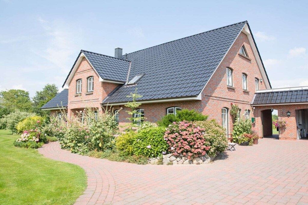 Een groot, met baksteen gebouwd huis met een steile dakschild, een verharde oprit en een verzorgde tuin met diverse planten en bloemen.