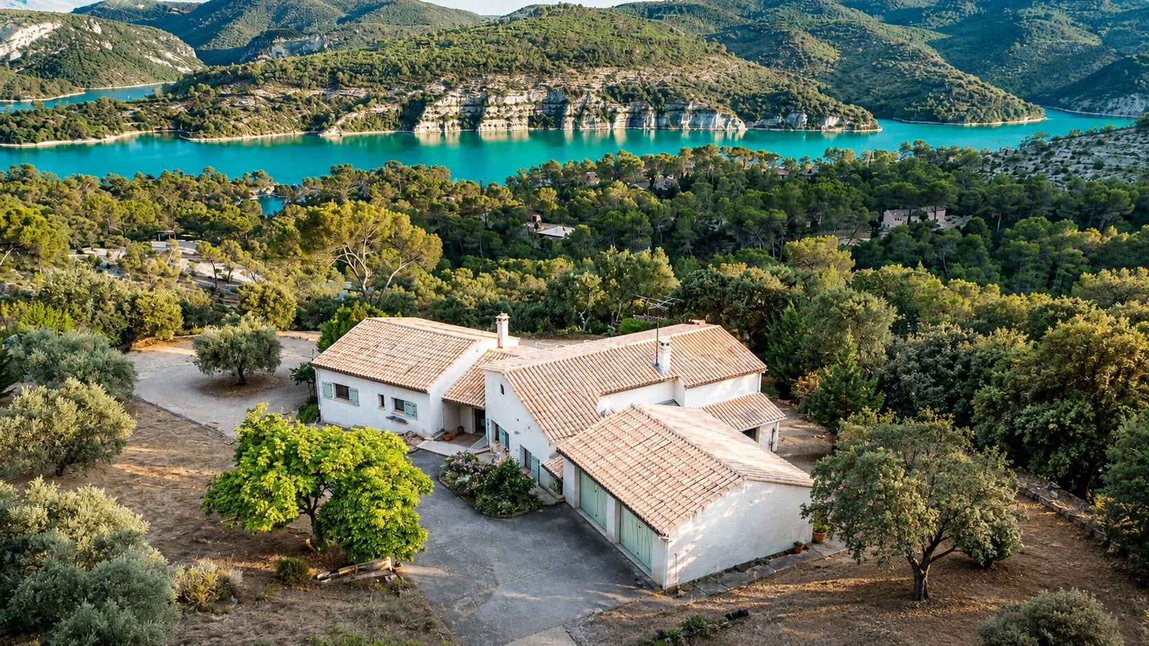 Un bâtiment blanc, doté d’un toit recouvert de tuiles, entouré d’arbres et bordé par un lac.