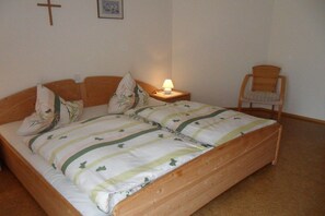 A single bed with a wooden frame, a bedside table with a lamp, a chair, and a cross on the wall. - Holiday farm Kleeblatt (Westheim)