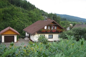 Ein zweistöckiges Haus mit geneigtem Dach, einer kleinen angebauten Garage und einem Balkon mit Blumen.