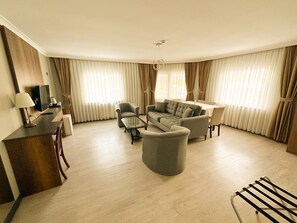 Amplia sala de estar con sofá, sillones, una mesa de centro y televisor. Cuenta con un comedor con mesa y sillas. La habitación tiene ventanas grandes con cortinas.