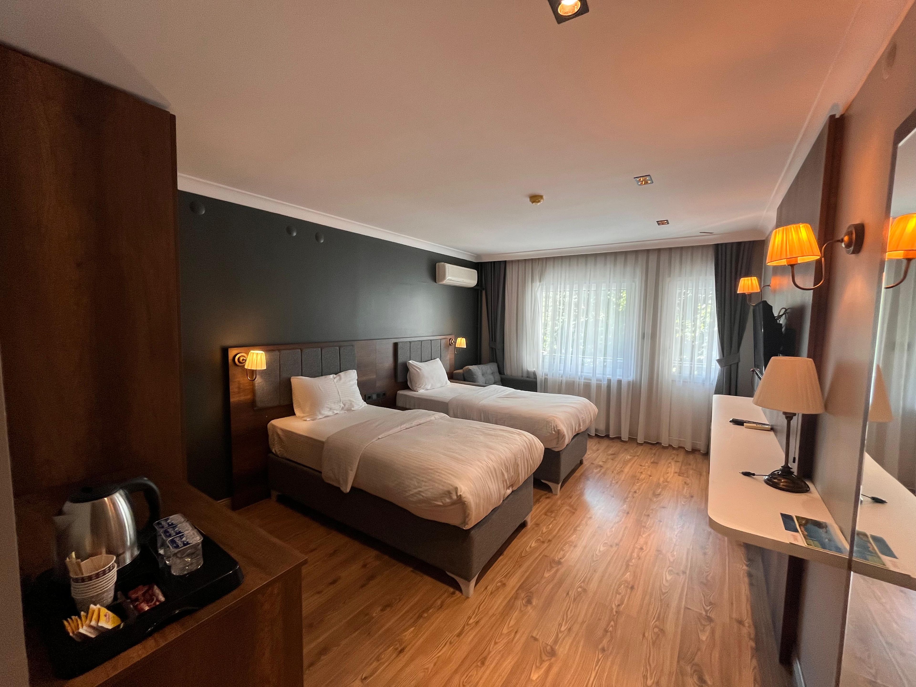 Habitación de hotel con dos camas, una mesita de noche con hervidor y vasos, un escritorio pequeño con lámpara y una ventana con cortinas.
