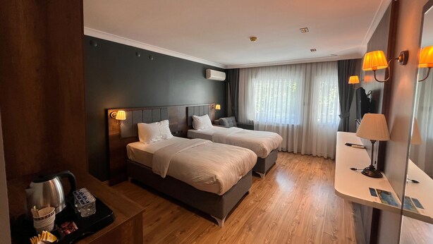 A hotel room with two beds, a bedside table with a kettle and glasses, a small desk with a lamp, and a window with curtains. - GRAND PALACE HOTEL (Bartın)