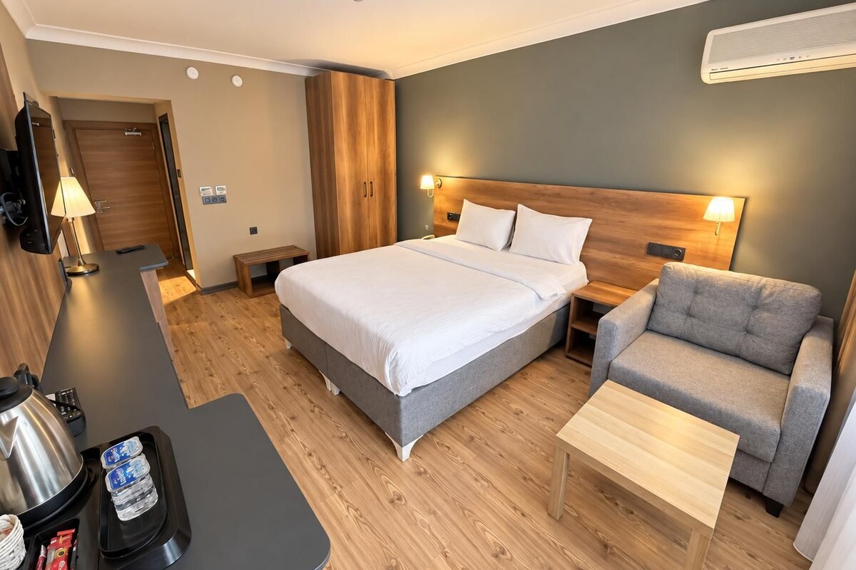 A hotel room with a bed, a sofa, a small table, a desk with a kettle, and a TV.