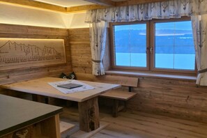 A wooden cabin interior with a dining table, benches, a window with curtains, and a wall-mounted light.