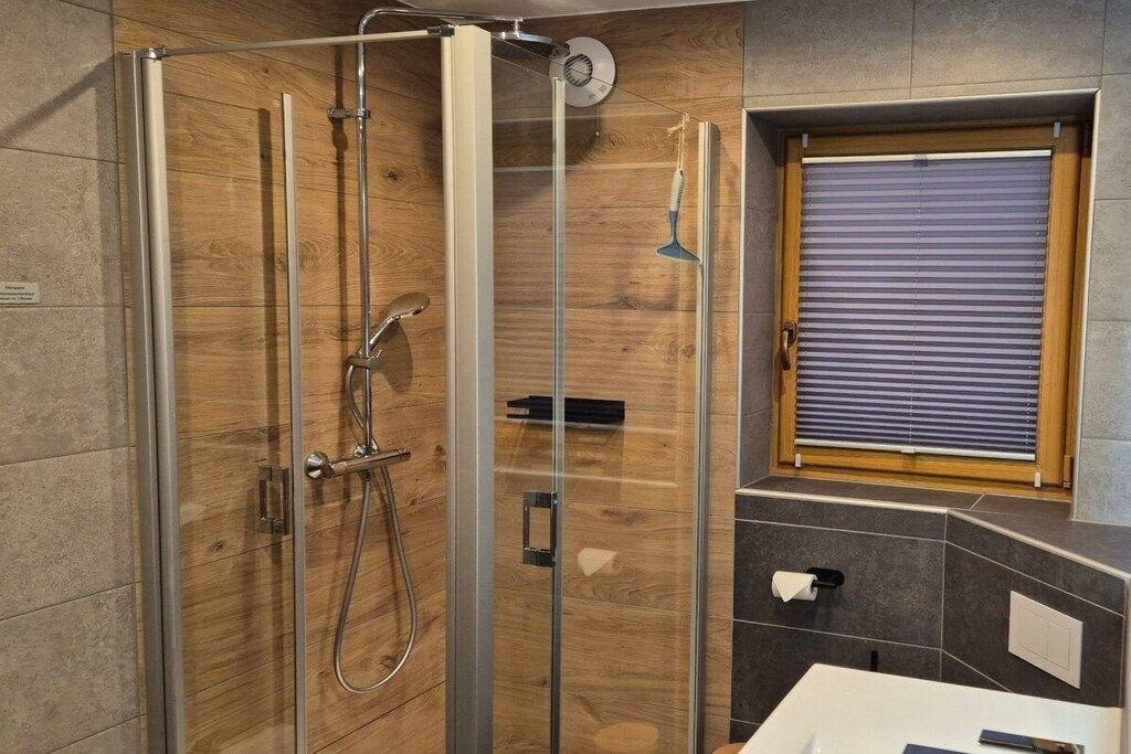 Ein modernes Badezimmer mit einer gläsernen Duschkabine, einem Holzbadezimmer-Möbel und einem Fenster mit Jalousien.