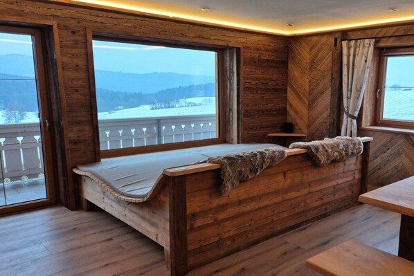 A wooden cabin with a bench and a table, large windows, and a view of a snowy landscape.