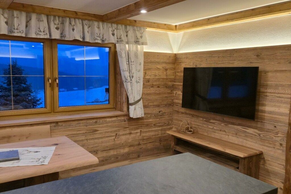 A cozy room with a wooden interior, a large window with curtains, a flat-screen TV mounted on the wall, and a bench.