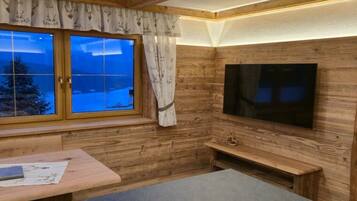 A cozy room with a wooden interior, a large window with curtains, a flat-screen TV mounted on the wall, and a bench.