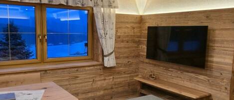 A cozy room with a wooden interior, a large window with curtains, a flat-screen TV mounted on the wall, and a bench.
