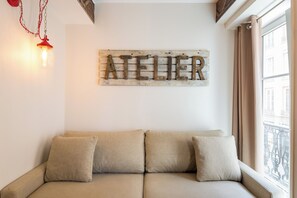 Un sofá de color neutro con tres cojines, un letrero de madera con la palabra 'ATELIER' en letras metálicas y una lámpara colgante roja.