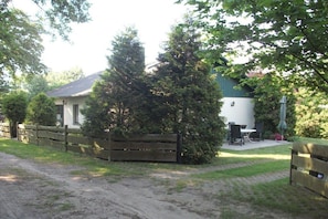 Ein eingezäunter Außenbereich mit Tisch und Stühlen, ein Gebäude mit weißer Fassade und ein Feldweg, der zum Eingang führt.