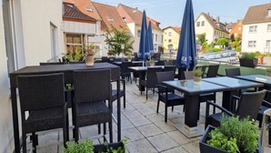 Ein Terrassenbereich im Freien mit Tischen, Stühlen, Blumentöpfen und blauen Sonnenschirmen.
