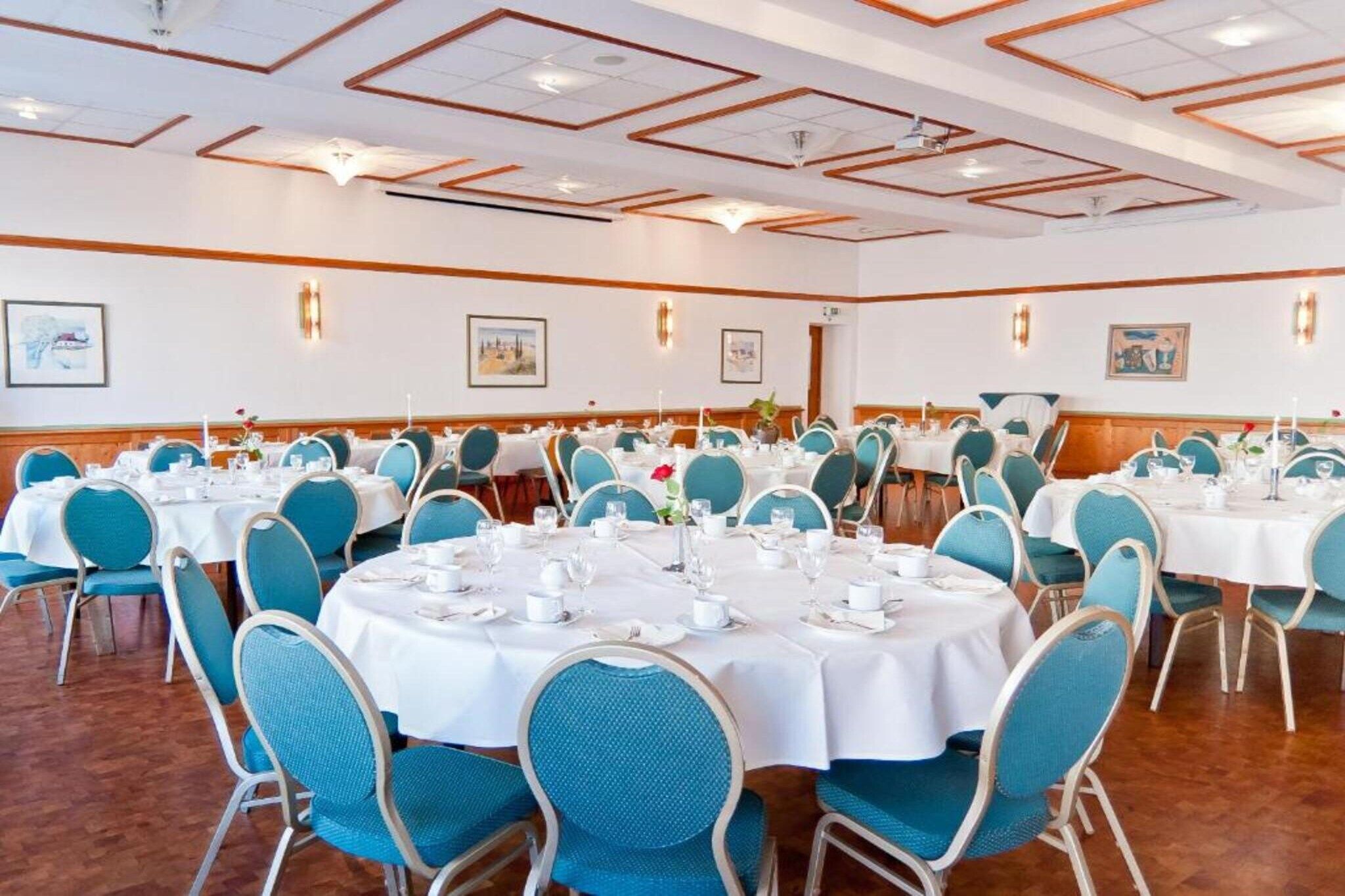 A banquet hall with round tables, blue chairs, white tablecloths, and framed pictures on the walls.