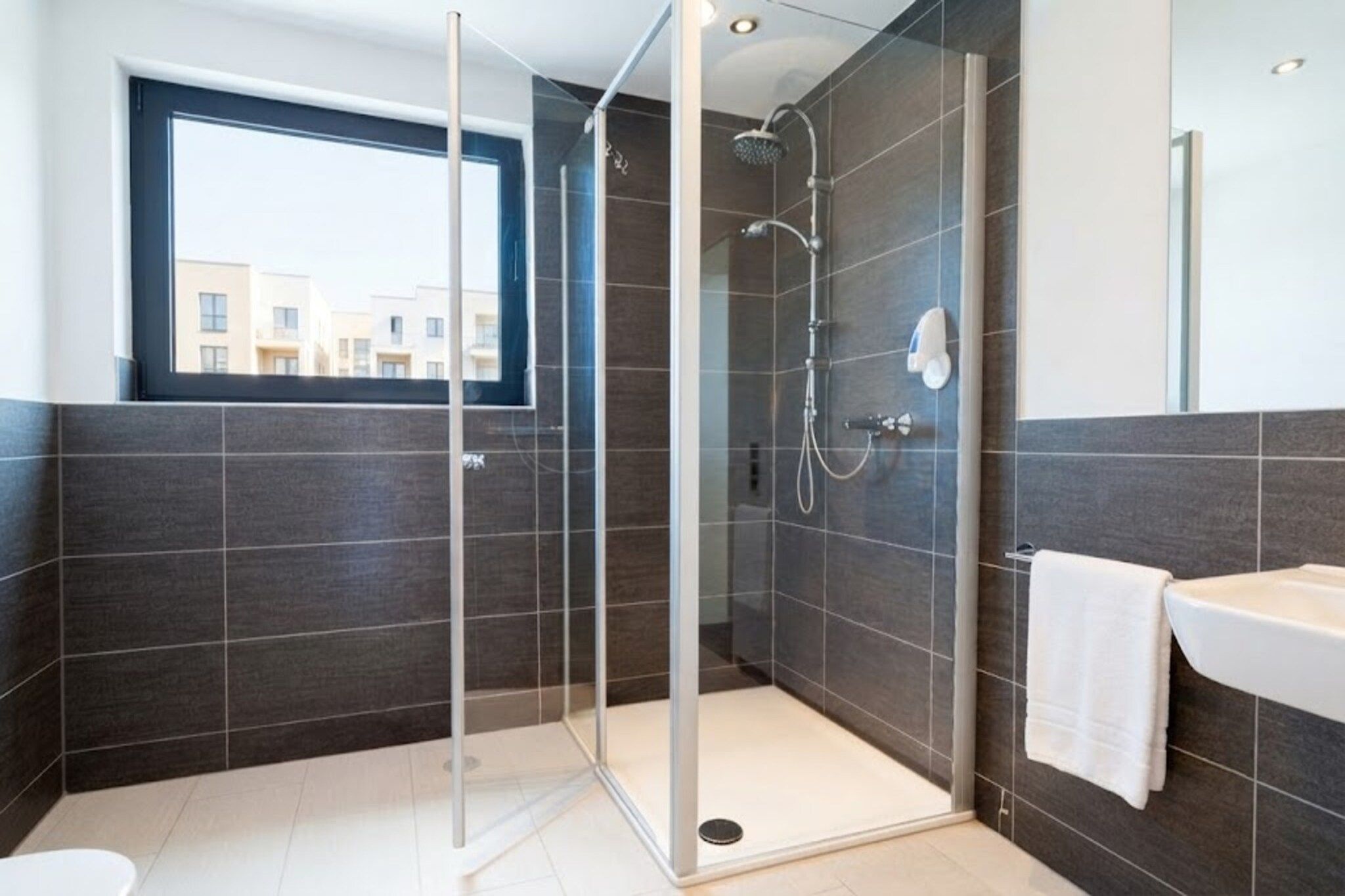 Un baño moderno con ducha acristalada, lavabo blanco y ventana con vistas a edificios.