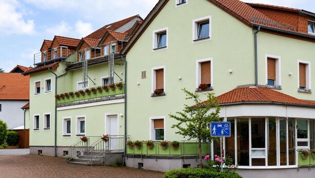 Ein mehrgeschossiges Gebäude mit grüner Fassade, einem überdachten Eingang und einem blauen Schild, das eine Einrichtung für Menschen mit Behinderungen kennzeichnet.